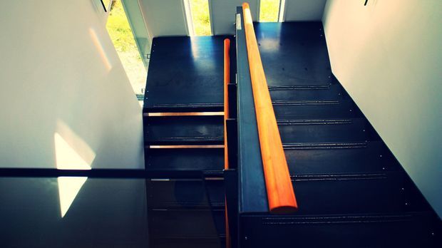 A dramatic interior view looking down a modern staircase with dark-painted stair treads and a contrasting warm timber handrail. Tall glazing at the landing brings in bright natural light and views of greenery outside, while strong geometric shadows fall across the pale wall.