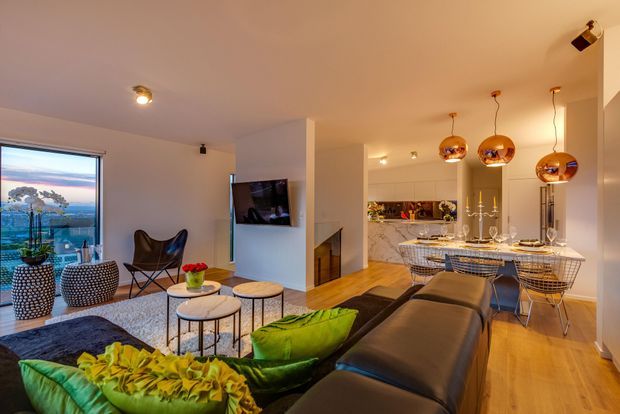 Open-plan contemporary living and dining area with a kitchen at the rear. The room features warm timber flooring, a large dark sectional sofa, bright green accent cushions, a textured dark rug, marble-look kitchen surfaces, wire-frame dining chairs, and three copper pendant lights over the dining island. Floor-to-ceiling glazing at left provides a broad view outside.