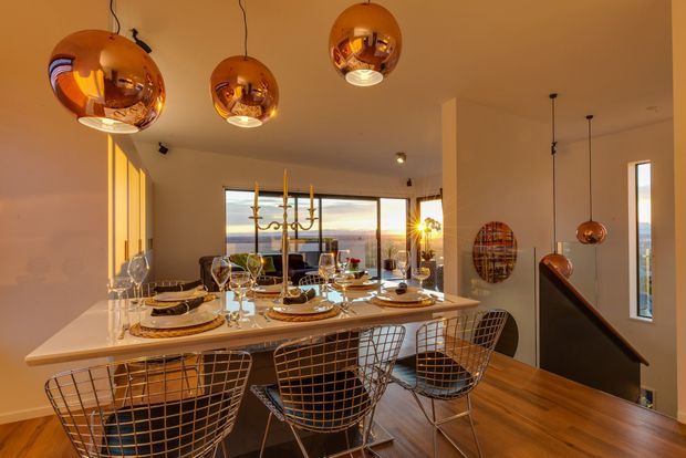 A warm, polished open-plan dining and kitchen space photographed at dusk. A white kitchen island or dining counter is set for a formal meal with layered plates, glassware, cutlery and tall candles, surrounded by wire-frame bar stools. Several large copper-toned spherical pendant lights hang overhead. Timber flooring, pale walls, expansive glazing with a distant view, and a stair opening with a dark handrail complete the scene.