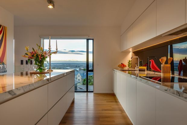 A refined modern kitchen viewed toward a glazed balcony or window with expansive water and landscape views. The room features two long runs of white handleless cabinetry, a large central island with a pale veined stone benchtop, a dark reflective splashback, warm timber flooring, under-cabinet lighting, and contemporary pendant and track lighting. A dining or living area is suggested beyond the island, with artwork, flowers, and decorative objects adding colour.