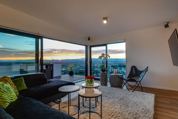 A contemporary living room with expansive corner glazing overlooking a broad landscape and sunset. The room features a dark sectional sofa, round nesting coffee tables, a textured light rug, a black leather sling chair, patterned ottomans, indoor plants, recessed and track lighting, and warm timber flooring. The open glazing creates an airy, panoramic outlook.