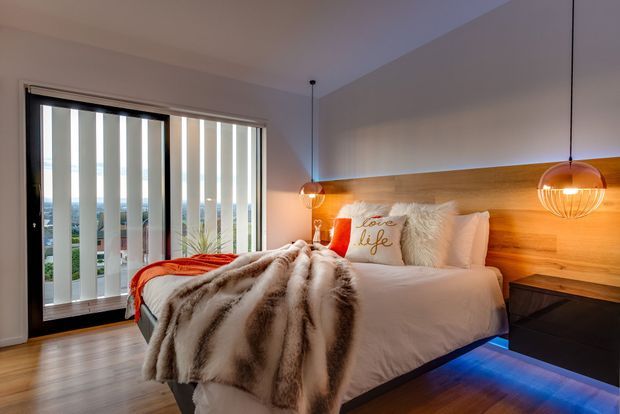 A warmly lit contemporary bedroom with a bed positioned against a full-width timber feature wall and integrated warm LED lighting beneath it. The room includes pendant lights, black bedside furniture, layered white bedding, a plush faux-fur throw, and orange accent cushions. Full-height vertical blinds open to a glazed balcony or exterior view, while timber flooring and neutral walls complete the space.
