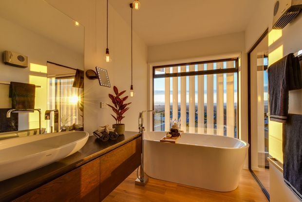 Warmly lit contemporary bathroom featuring a freestanding white bathtub beside floor-to-ceiling glazing with vertical blinds, a timber vanity with a white vessel basin, large wall mirror, pendant lights, and dark towel storage. The room has a calm, refined atmosphere with timber flooring and views beyond the windows.