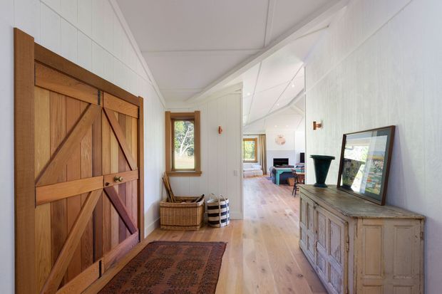 Bright farmhouse-style entry hallway with white vertical wall panelling, pale timber flooring, a large rustic timber barn door, and a distressed painted timber console. The corridor opens toward a furnished living area, with natural light entering through timber-framed windows and glazed openings.