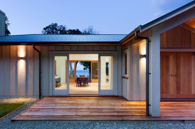 A dusk view of a contemporary single-storey courtyard-style home, focused on a timber deck and glazed entry opening. Vertical grey cladding, dark metal roofing and gutters, warm exterior wall lights, and timber-framed architectural elements surround a central glazed connection to an interior dining area. A timber-clad garage or ancillary volume is visible on the right.