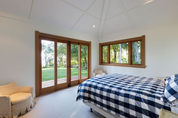 Bright bedroom with a vaulted white ceiling, warm timber-framed glazed doors and windows, pale carpet, and a bed dressed in navy-and-white checked bedding. A cream upholstered armchair sits near the glazed doors, which open toward a leafy lawn and garden.