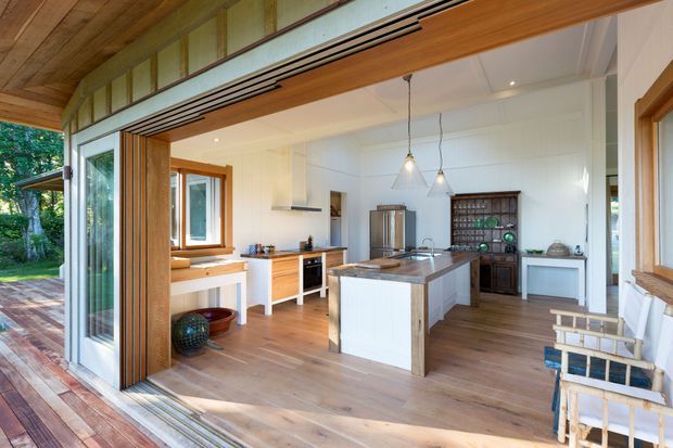 Bright open-plan kitchen viewed from a timber-framed indoor-outdoor opening, featuring warm timber flooring and ceiling boards, white paneled walls, a central island with a dark stone benchtop, white cabinetry, timber-framed glazing, and a built-in dark timber display cabinet. The room opens onto a deck or terrace with garden views.