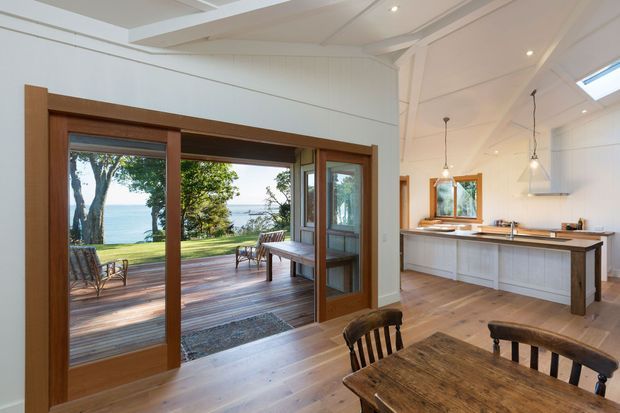 Bright open-plan coastal home interior with a dining area in the foreground and a white, timber-lined kitchen beyond. Wide timber-framed sliding doors open onto a spacious timber deck and lawn with a visible waterfront outlook. Exposed and painted timber ceiling beams, pale timber flooring, white cabinetry, and a long timber-topped island create a warm, relaxed setting.