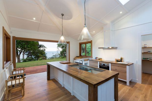 A bright, spacious coastal-style kitchen with a large timber-topped island, white panelled cabinetry, timber flooring, pendant lights, and stainless-steel appliances. Large openings connect the kitchen to a timber deck and leafy ocean-facing outlook, while a skylight and recessed ceiling lights add natural and artificial illumination.