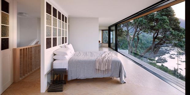 Bright contemporary bedroom with a low-profile bed, white bedding, and a pale throw positioned beside expansive floor-to-ceiling glazing overlooking mature trees. A timber balustrade and hallway are visible at left, while textured glass-panel doors add architectural detail.
