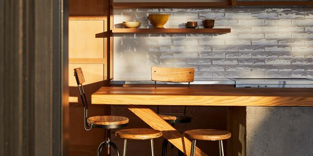 A warm, compact kitchen or dining counter with a thick timber benchtop, integrated stools, dark wood cabinetry, open shelving, and a textured grey-white tiled splashback. Strong sunlight creates a rich, mid-century-inspired atmosphere.