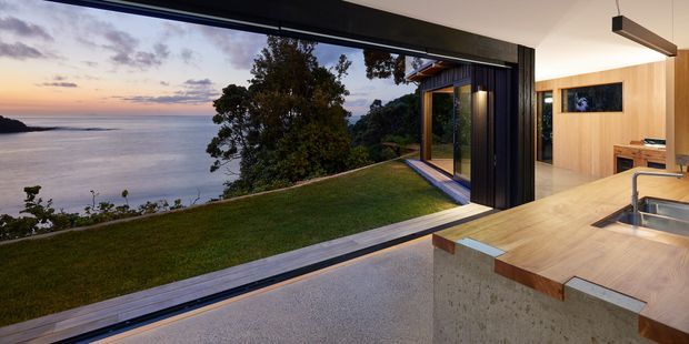 Open-plan contemporary kitchen and living space with a timber island and concrete base, opening through extensive floor-to-ceiling glazing to a manicured lawn and expansive waterfront view at dusk. Dark framed sliding doors, warm timber wall panelling, integrated cabinetry, and a linear pendant reinforce the refined coastal setting.