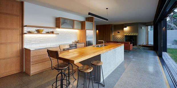 Open-plan contemporary kitchen and living area with a long timber island and integrated seating, pale stone or concrete island base, timber cabinetry, a textured light backsplash, polished concrete flooring, and extensive dark-framed glazing opening toward a small garden. A living area with warm timber wall panelling and a red sofa is visible beyond the kitchen.