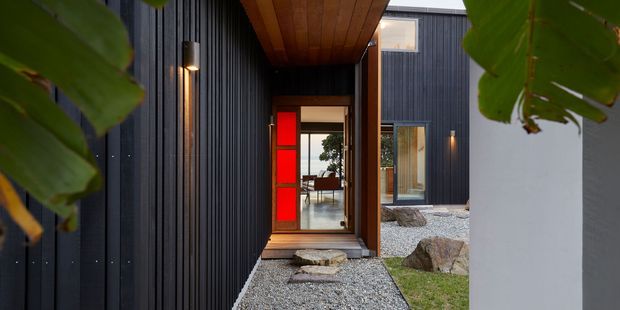 A modern residential entry approach framed by lush foliage, with dark vertically ribbed cladding, a warm timber soffit, a glazed front door with vivid red side panels, contemporary wall lights, and a minimalist gravel courtyard with natural stone accents.