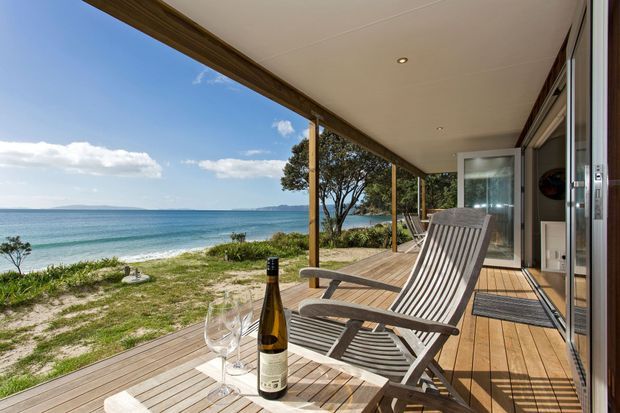 A relaxed beachfront deck overlooking the ocean, with timber decking, a covered roof, glass sliding doors, and a reclining outdoor chair beside a small table set with wine glasses and a bottle. The scene has expansive coastal views and a bright, tranquil atmosphere.
