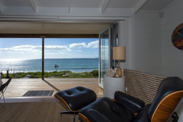 A coastal living room opens directly onto a timber deck through wide glazed folding doors, with expansive ocean views. The interior features a white painted ceiling with exposed beams, white wall panelling, dark leather lounge chairs and ottoman, a textured woven wall feature, and a warm timber-framed outlook.