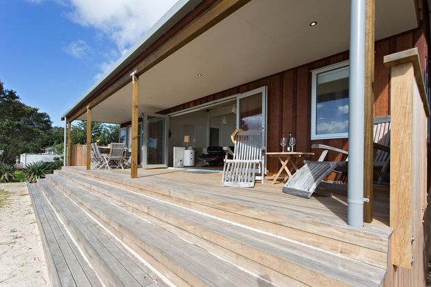 A single-storey timber-clad house with a broad covered verandah and expansive timber deck. Wide steps lead up to the outdoor living area, which has sliding glass doors, timber posts, outdoor chairs, a small table, and a partially visible interior. The setting appears sunny and relaxed, with landscaped vegetation at the edges.
