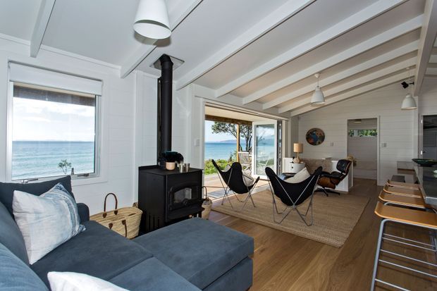 Bright coastal living space with white-painted timber walls and exposed ceiling beams, featuring a dark sectional sofa, black wood-burning stove, woven rug, and iconic black sling chairs. Large windows and glazed doors provide ocean views and open onto a timber deck. A kitchen island with bar stools is visible at right.