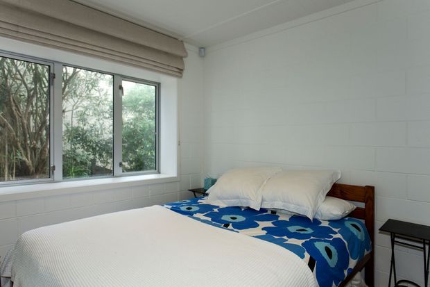 A compact, bright bedroom with a timber bed, white bedding, and a bold blue-and-white patterned bedspread. White painted horizontal wall panelling, a large window with a beige Roman blind, and simple black bedside furniture create a clean, relaxed interior overlooking greenery.