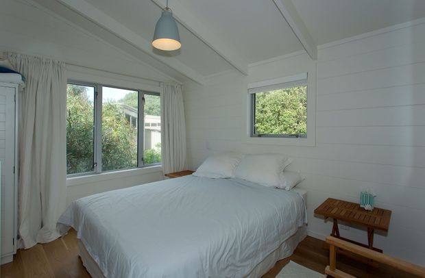 A bright, compact bedroom with a white bed, white bedding, white horizontal wall panelling, and timber flooring. Large windows with curtains bring in natural light and overlook greenery. A small timber bench and side table sit beside the bed, with a simple pendant light overhead.