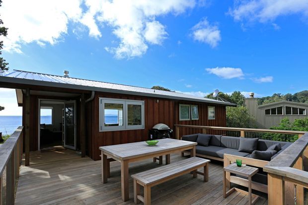 A timber-clad coastal deck and outdoor entertaining area beside a compact single-storey dwelling. The space features a large timber dining table with benches, an outdoor sectional lounge, coffee tables, timber decking and railings, and a low-pitched metal roof. Ocean water is visible beyond the building, with neighbouring structures and vegetation in the background.