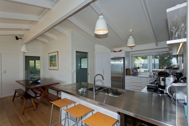 Bright open-plan kitchen and dining area in a white-lined interior, featuring a large stainless-steel island with integrated sinks, timber bar stools, white cabinetry, stainless appliances, pendant lighting, exposed ceiling beams, and an adjoining timber dining table.