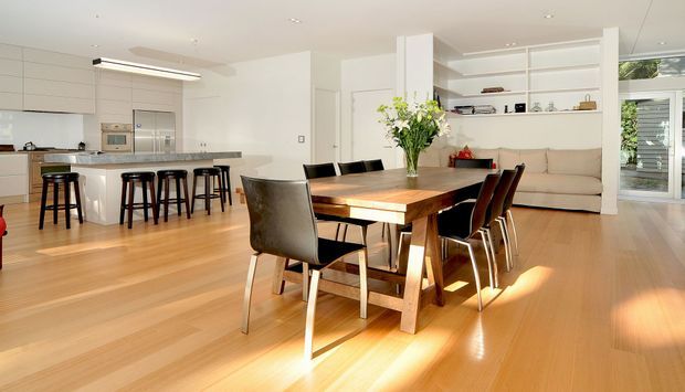 Bright open-plan interior featuring a contemporary white kitchen with a long island and bar seating, opening into a dining area with a substantial timber dining table and dark upholstered chairs. Built-in white shelving and a pale sofa sit along the rear wall, with light timber flooring, recessed lighting, and abundant natural light from the glazing on the right.