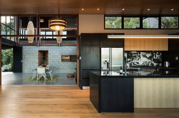 A contemporary open-plan kitchen and dining area with dark timber cabinetry, a central island, stainless-steel appliances, and a black-and-white patterned splashback. Warm timber flooring and a timber-lined ceiling contrast with pale masonry walls in the adjacent dining zone. Large glazed openings, clerestory windows, pendant lights, and an internal balcony/void contribute to the airy, architecturally detailed space.