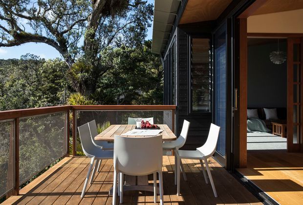 A timber outdoor dining deck adjoining a contemporary home, with a rectangular timber table and white chairs enclosed by a transparent balustrade. Large glass sliding doors open into a dark-clad interior, where a bedroom is visible. The deck is surrounded by mature trees and dense greenery, creating a private elevated setting. Strong natural daylight contrasts warm timber surfaces with dark exterior cladding and white furniture.
