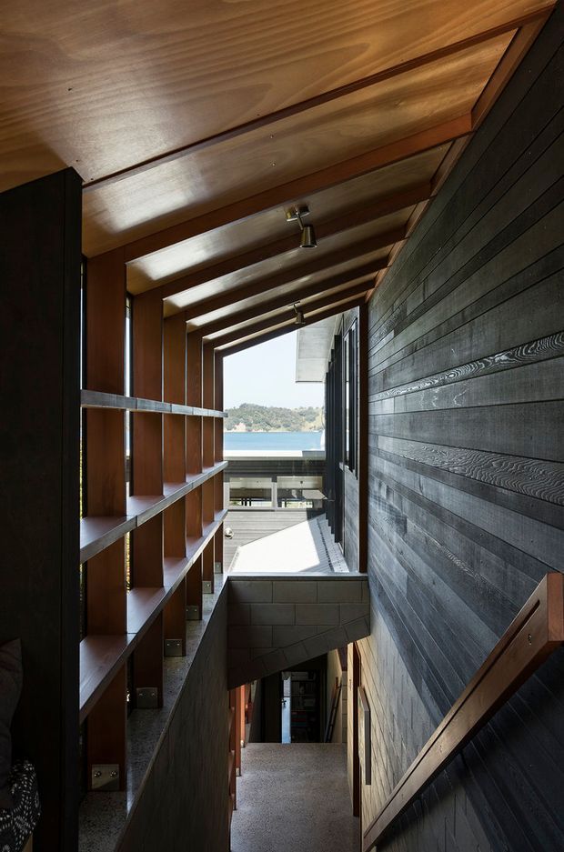 A narrow, dramatic interior stairway and landing with a long view toward a bright waterfront or coastal landscape. The space features exposed warm timber roof beams and ceiling boards, dark horizontal timber wall cladding, open timber shelving, concrete blockwork, and a timber handrail. Small recessed or surface-mounted spotlights run along the ceiling, while strong daylight enters through the open end and vertical glazing.