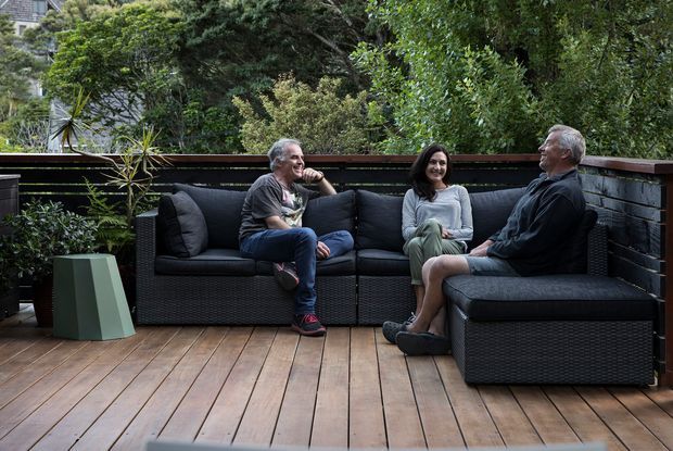A landscaped outdoor terrace with a warm timber deck, dark horizontal screening and a low railing, furnished with a large dark woven sectional sofa and scattered cushions. Three adults are seated in conversation, surrounded by dense greenery and potted plants. The setting feels private, relaxed and sheltered, with a muted contemporary outdoor-living aesthetic.