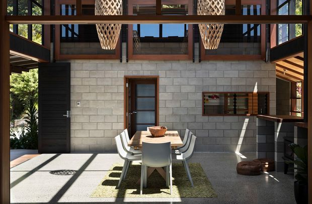 A bright, double-height dining and entertaining space with exposed dark timber framing, large windows, and a light grey concrete block feature wall. A central dining table with modern white chairs sits on a textured rug, while a compact bar area occupies the right side. Woven pendant lights hang overhead, and strong sunlight creates geometric shadows across the polished concrete floor.