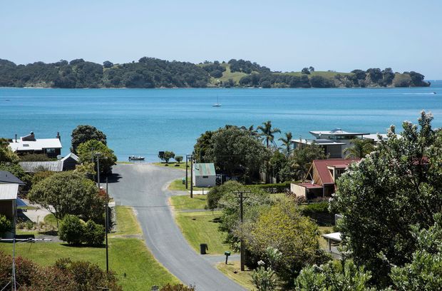 A bright elevated view over a quiet coastal residential settlement, with a paved road descending toward vivid turquoise water and a wooded, rolling shoreline across the bay. Low-rise detached homes, pitched roofs, gardens, utility poles, and a small sailboat are visible.