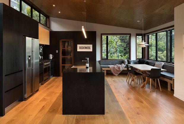 Open-plan kitchen and dining area with a dark, monolithic kitchen island and matching full-height cabinetry, integrated stainless-steel appliances, warm timber flooring, and a wood-panelled ceiling. A dining table with black chairs sits beside built-in window seating, while extensive glazing provides views of greenery. The space has a refined, contemporary feel with strong natural materials and warm ambient lighting.
