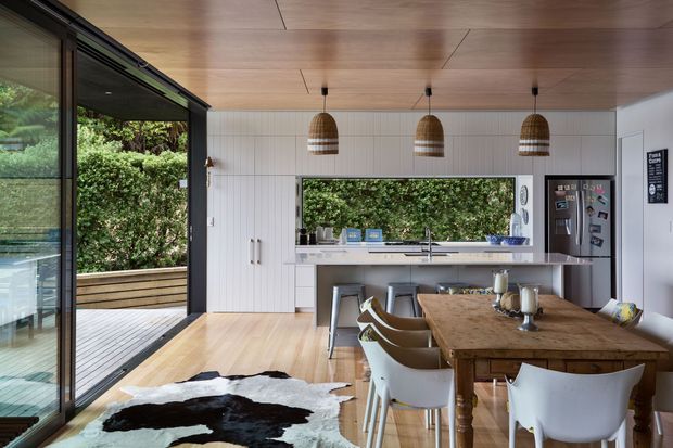 A bright, open-plan dining and kitchen interior with large sliding glass doors opening to a timber deck and lush greenery. The kitchen features white vertical-panel cabinetry, a long island with a pale benchtop, and three woven pendant lights. A substantial timber dining table with modern white chairs occupies the foreground, while warm timber flooring and ceiling lining create a cohesive natural palette.