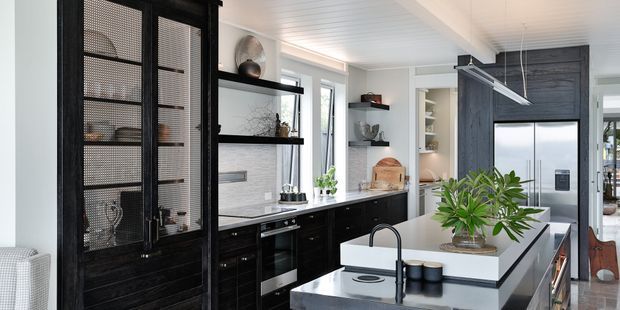 A bright contemporary kitchen with black built-in cabinetry, a tall mesh-front display cabinet, white tiled or panelled walls, stainless-steel appliances, and a large central island with a pale stone benchtop. Open shelving, integrated lighting, indoor plants, and a white-lined ceiling create a refined monochrome interior.