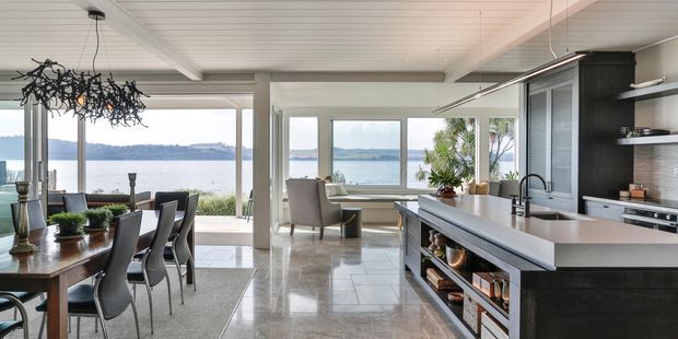 A bright, open-plan kitchen, dining, and living area with expansive glazing overlooking a body of water. The room features a large dark kitchen island with integrated storage, white cabinetry, polished light stone flooring, a timber dining table, contemporary dark dining chairs, and a sculptural branch-like pendant light. A sitting area is positioned by the windows, creating an airy waterfront-facing interior.