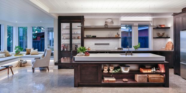 A bright, open-plan kitchen with a large central island, dark timber cabinetry, integrated shelving, and a pale stone benchtop. The kitchen opens directly to a light-filled living area with expansive windows. White painted ceiling boards, grey stone flooring, black-framed glazing, and warm styling create a refined contemporary interior.