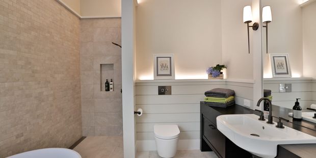 A bright, refined bathroom featuring a large walk-in shower clad in pale textured stone-look tile, a freestanding bathtub partially visible in the foreground, and a vanity with a white integrated basin, dark cabinetry, and black tapware. Horizontal painted wall panelling, framed artwork, integrated shelf lighting, and twin wall sconces add a polished contemporary-traditional character.
