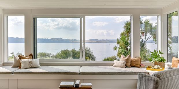 Bright, serene waterfront living space featuring a built-in window seat beneath expansive panoramic glazing. The room has pale neutral finishes, a white painted slatted ceiling, soft upholstered seating, timber-toned accents, and indoor plants, with a calm bay or lake view beyond.
