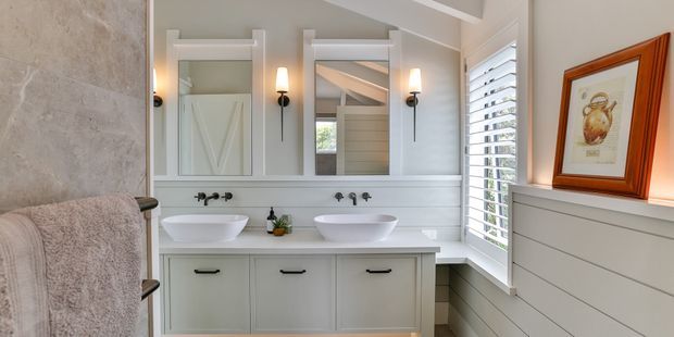 Bright, refined bathroom with a double vanity, two white vessel basins, pale grey cabinetry, marble-look wall finish, framed mirrors, wall sconces, and white plantation shutters. Horizontal wall panelling and warm timber-framed artwork add character.