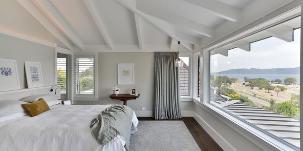 Bright coastal bedroom with a white upholstered bed, neutral bedding, framed artwork, a dark timber console table, grey carpet, and white-painted exposed ceiling beams. Large corner windows provide expansive views toward the water and surrounding landscape, while full-height striped curtains add texture.