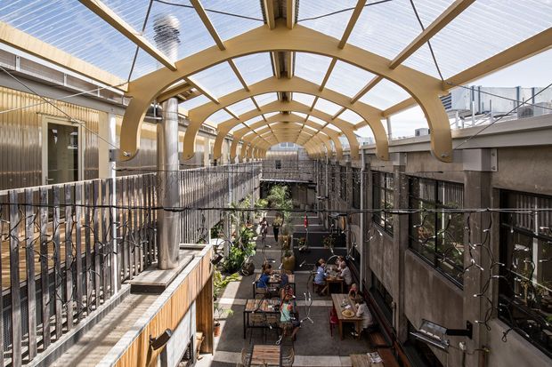 A covered multi-level commercial courtyard or laneway with a long translucent roof, repeated curved timber arches, exposed structural framing, metal railings, and dining tables below. Restaurants or hospitality spaces line both sides, with planting and string lights adding detail. The image is bright and airy, viewed from an elevated walkway toward the central communal area.