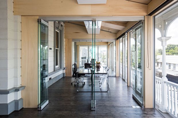 A bright enclosed verandah or glazed extension used as a home office, with a long desk and office chairs arranged along the space. The room has extensive floor-to-ceiling glazing and folding glass doors, dark timber flooring, pale timber ceiling and structural framing, skylights, and views onto a traditional white verandah. A white weatherboard wall and classical exterior detailing are visible at the left.