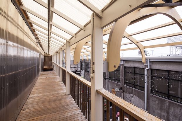 A covered elevated timber walkway or balcony runs along the side of a building. The space features timber decking and handrails, cream-painted structural posts and beams, translucent roof panels providing abundant daylight, and distinctive curved timber brackets. Corrugated translucent wall panels line the left side, while an adjacent lower-level courtyard or building facade is visible on the right.