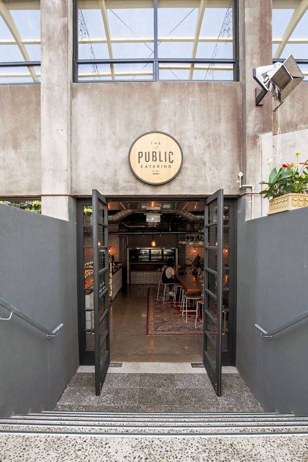 A downward-looking view into the entrance of The Public Catering Co., a commercial café or restaurant. Open black-framed glazed doors lead down a stairway into an industrial-style dining and bar area, with exposed ductwork, pendant lights, stools, tables and a patterned rug. The entrance is framed by raw textured concrete walls and columns, with a large glazed roof or clerestory above and dark grey stair walls with handrails. A planter is visible at the upper right.