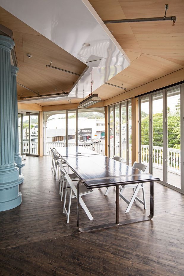 Bright, elongated dining or communal meeting space with a long dark table and white folding chairs. The room features extensive floor-to-ceiling glazed doors and windows opening onto a veranda, a warm timber-lined ceiling and dark timber flooring, exposed black pipework, industrial-style suspended lighting, and a large glossy white overhead ceiling bulkhead. A substantial fluted blue-green column anchors the left side, with an urban streetscape visible outside.