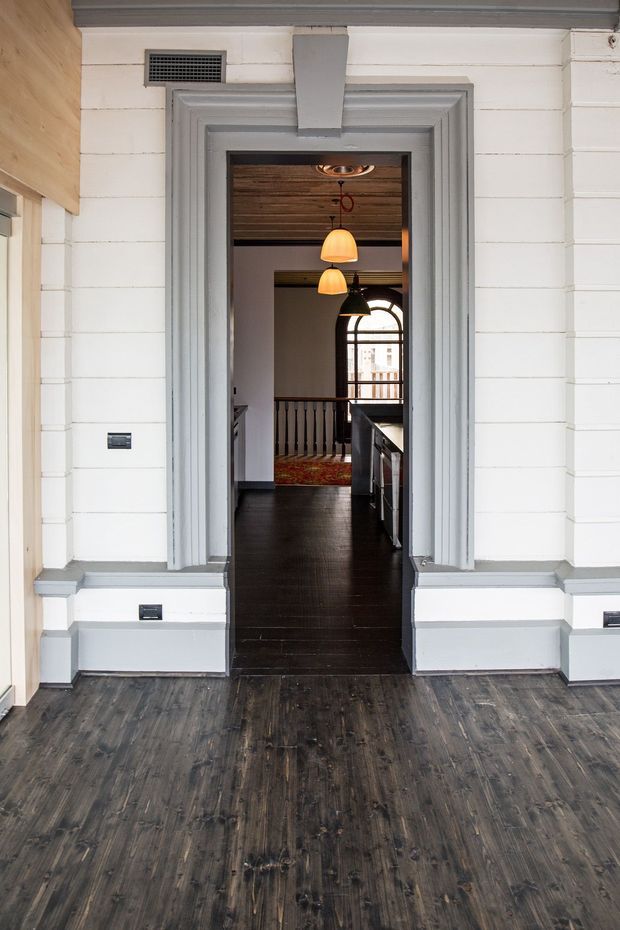 Interior view looking through an ornate grey-trimmed doorway into a hallway and adjoining kitchen area. The space features dark timber flooring, white horizontal wall panelling with substantial decorative skirting and architraves, a dark timber-plank ceiling beyond the doorway, warm pendant lights, a stair balustrade, and an arched window at the rear. The composition emphasises the traditional architectural detailing and layered rooms.