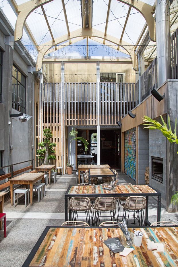 A bright, narrow urban hospitality courtyard or café dining space enclosed by a translucent glazed roof. The space features multiple timber dining tables with woven chairs and benches, a tall timber slatted screen, exposed steel and timber structural elements, concrete block walls, wall-mounted lighting, decorative string lights, potted greenery, and a built-in fireplace or heater along the right wall. The architecture combines an industrial shell with warm timber detailing and an airy atrium-like roof.