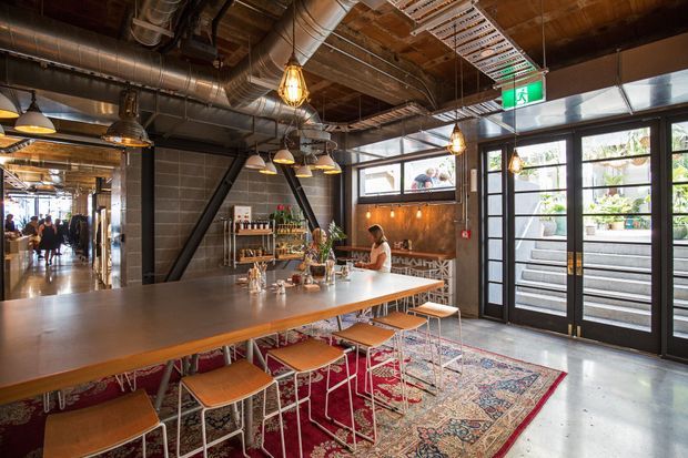An industrial-style commercial dining and bar space with a long communal timber-topped table and stools in the foreground. The room features exposed timber ceiling structure, visible metal ductwork, suspended pendant lights, dark steel structural bracing, concrete flooring, and large black-framed glazed doors opening to an outdoor area. A bar or service counter, shelving, plants, and patrons are visible toward the rear.